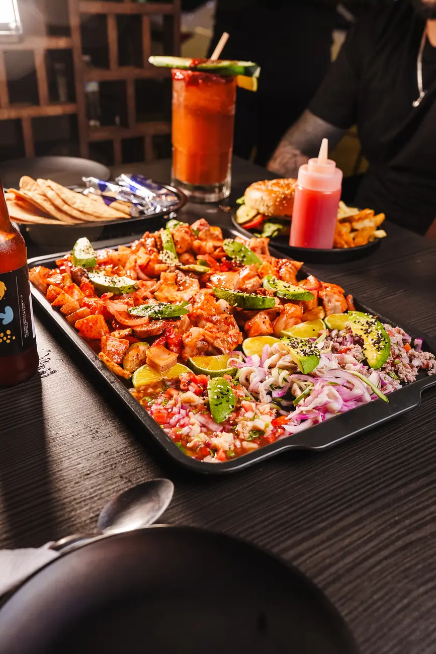 Charola de mariscos y cerdo con aguacate y limón sobre la mesa de Latino, con una michelada y una hamburguesa al fondo