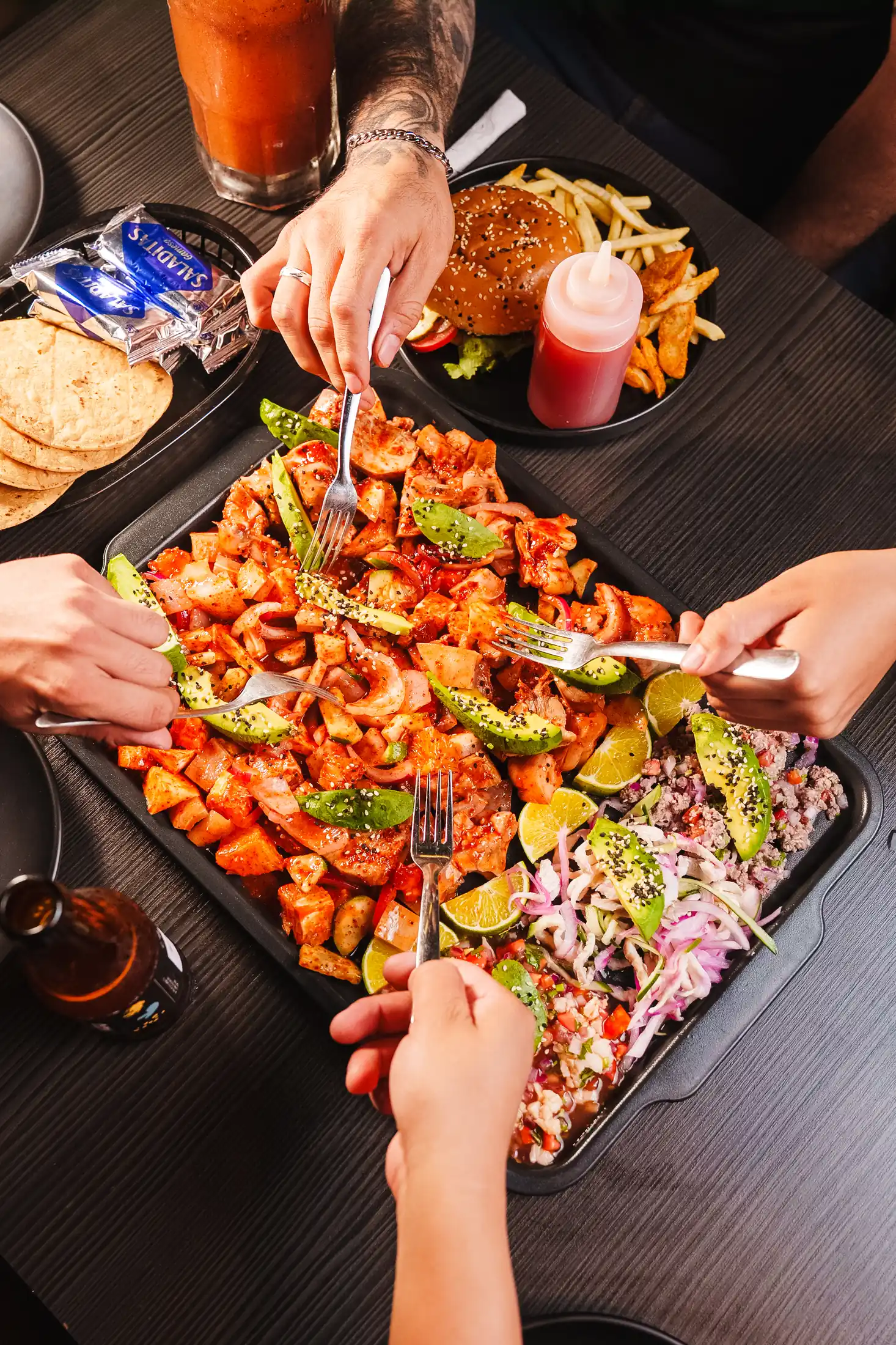 Vista cenital de una charola compartida entre cuatro manos, con hamburguesa, papas, michelada y tostadas alrededor
