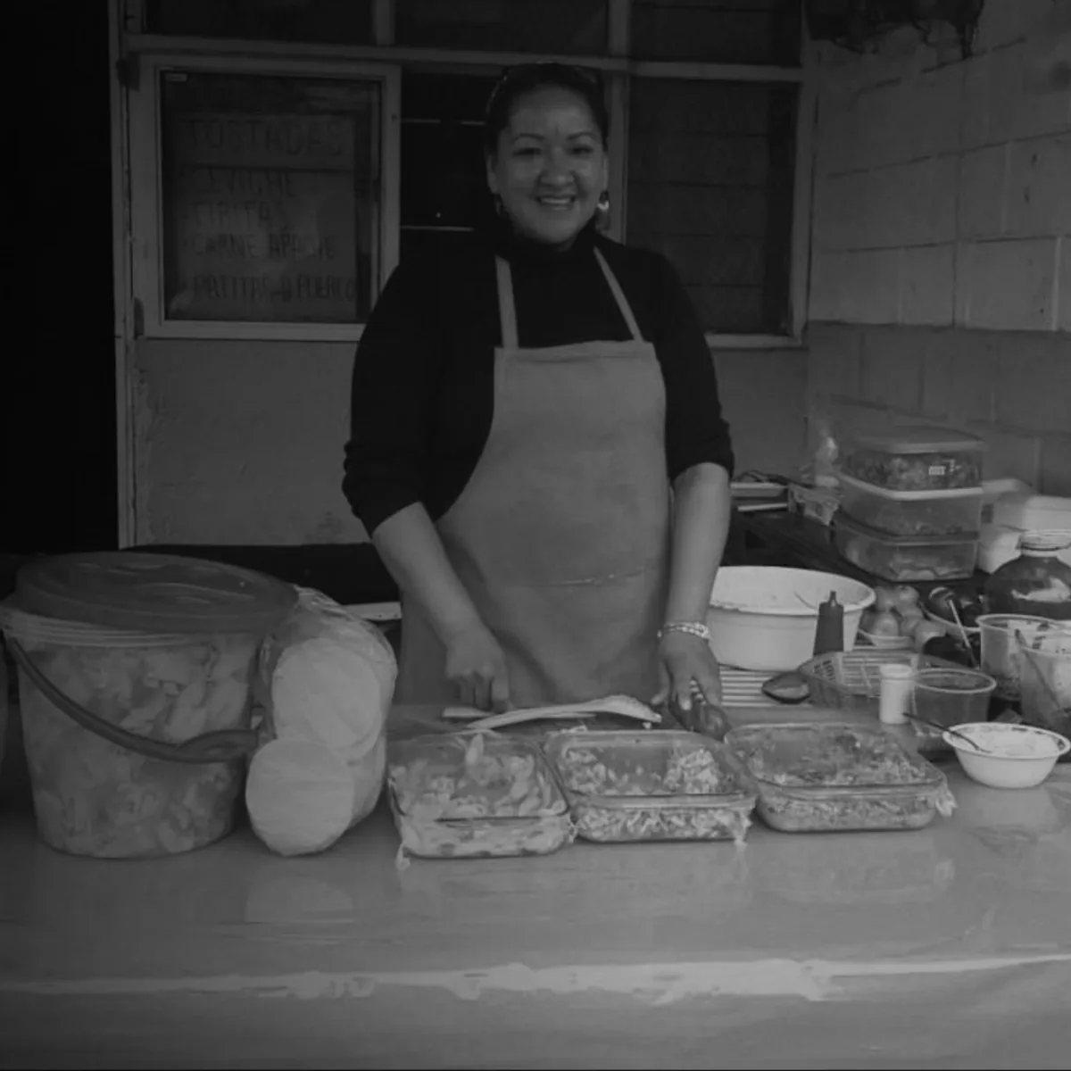 Fotografía en blanco y negro de una mujer sonriente detrás de un puesto callejero, con charolas de mariscos preparados y un menú escrito a mano al fondo
