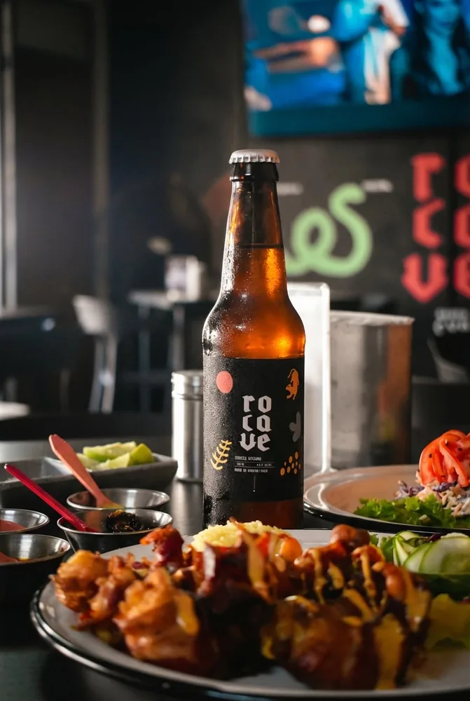 Foto real: una Rocave de pie sobre la mesa con salsas y platos alrededor, mural de Rocave desenfocado al fondo